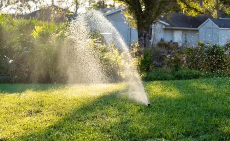 Sprinkler watering a sunlit, lush green lawn in a garden. Water droplets sparkle in the sunlight