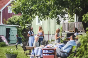 A backyard barbecue with several people socializing and dining at a table with striped tablecloths